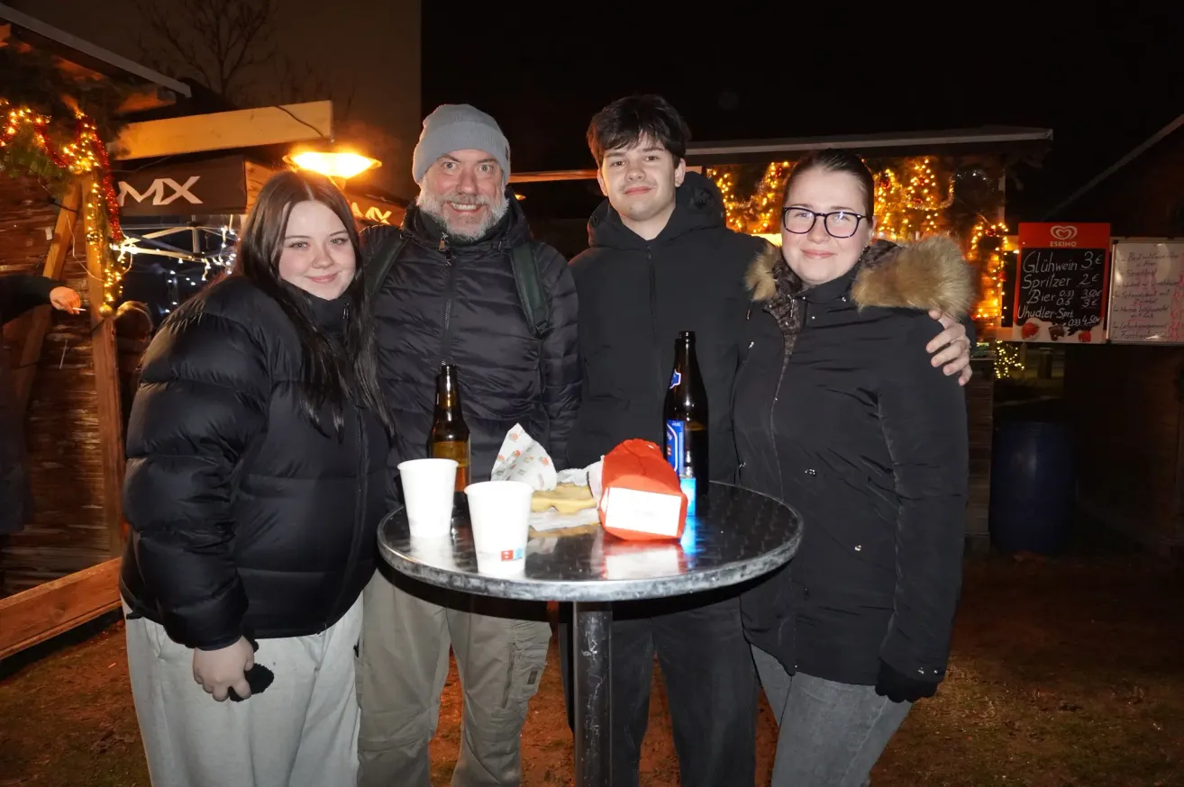 Vier Menschen stehen in einer Nachtaufnahme zusammen und lächeln. Vor ihnen stehen auf einem Tisch zwei Gläser, zwei Flaschen und eine Tasche.