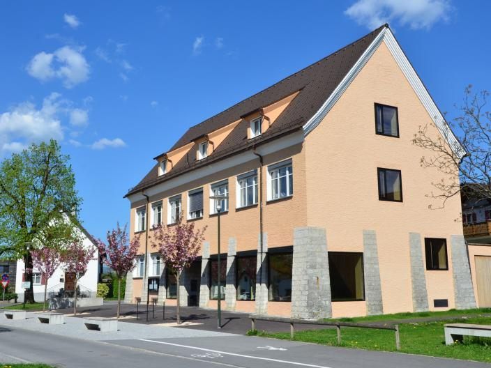Ein dreistöckiges Gebäude mit einem braunen Dach steht an einer Straße, umgeben von Bäumen und Gras. Die Fassade weist zahlreiche Fenster auf, und eine Straßenlaterne ist davor positioniert.