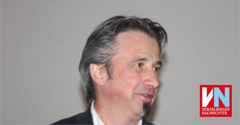 Ein Mann mit grauem Haar und einem Schnurrbart trägt einen schwarzen Anzug. Er schaut nach rechts und lächelt.