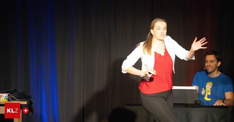 Eine Frau in einem roten Oberteil und schwarzen Hosen steht auf der Bühne. Sie trägt eine weiße Jacke und hält ein rotes Objekt.