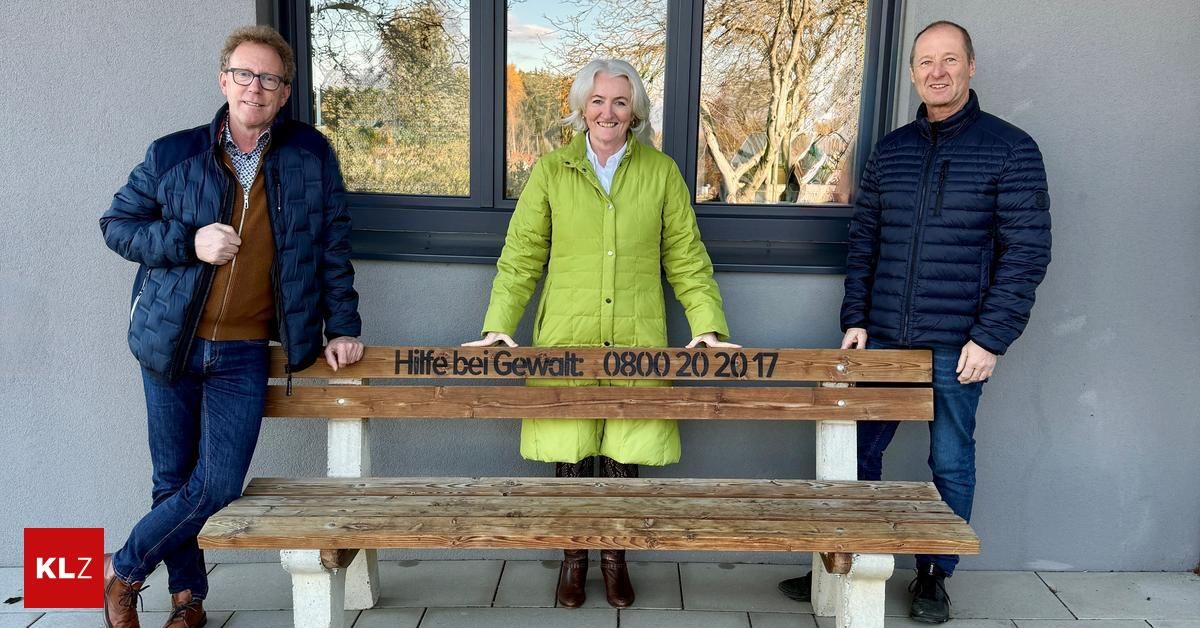 Drei Menschen posieren vor einer Bank. Die Frau in der Mitte lächelt. Die Bank hat einen Spruch, der lautet: 'Hilfe bei Gewalt: 0800 20 20 17.'