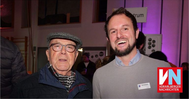 Zwei Männer stehen bei einer Veranstaltung zusammen. Der Mann auf der linken Seite trägt eine Mütze, eine Brille und einen Schal. Der Mann auf der rechten Seite hat einen Namensschild. Hinter ihnen stehen und unterhalten sich Leute.