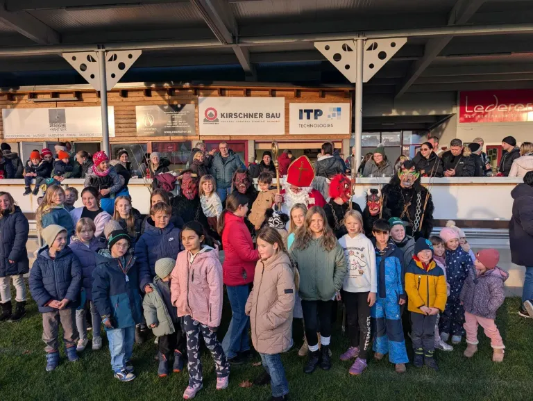 Eine Gruppe von Kindern und Erwachsenen, einige in Kostümen gekleidet, posiert für ein Foto vor einem Gebäude mit Schildern für Kirschner Bau und ITP Technologies.