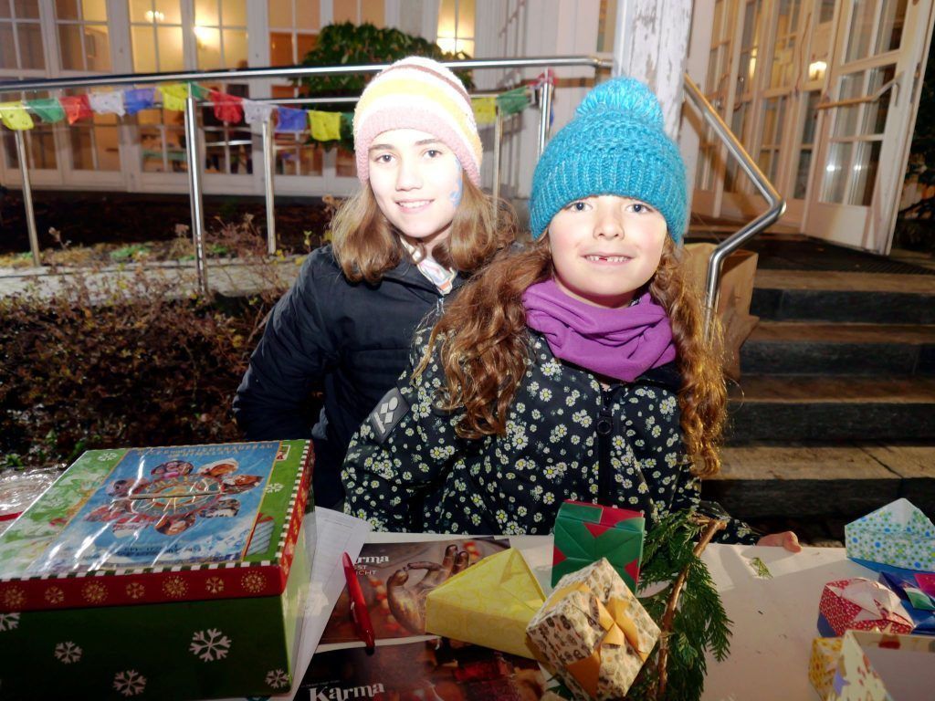 Zwei junge Mädchen sitzen an einem Tisch mit Weihnachtsdekoration. Das Mädchen auf der rechten Seite hat Gesichtsbemalung und lächelt. Auf dem Tisch liegen Schachteln, Geschenke und ein Stift. Dahinter befindet sich eine Treppe mit Geländer.