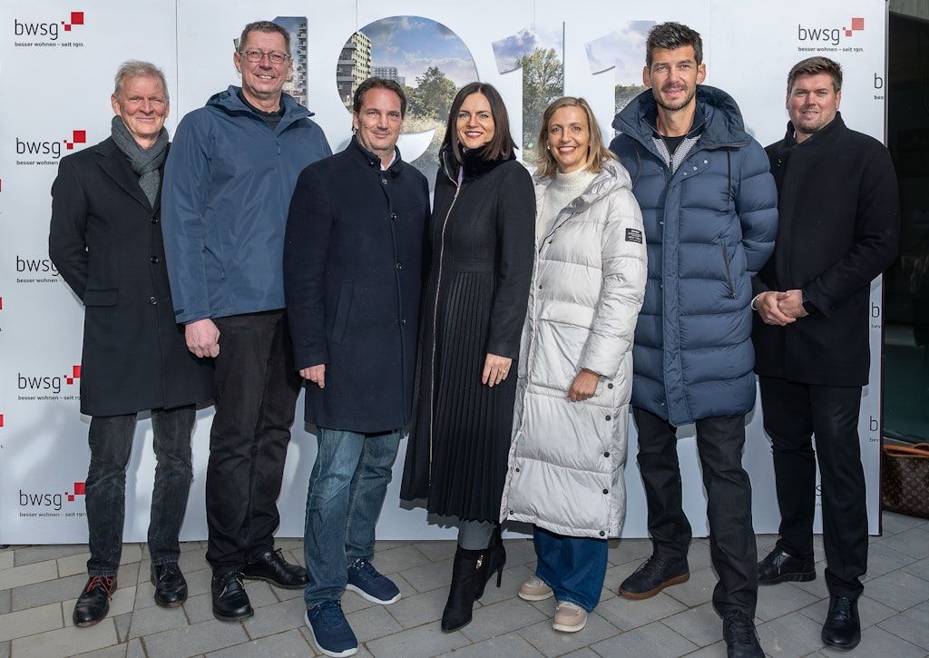 Eine Gruppe von sieben Personen steht eng beieinander vor einer weißen Wand und trägt Winterkleidung. Der Mann in der Mitte trägt eine Brille und einen Ring.