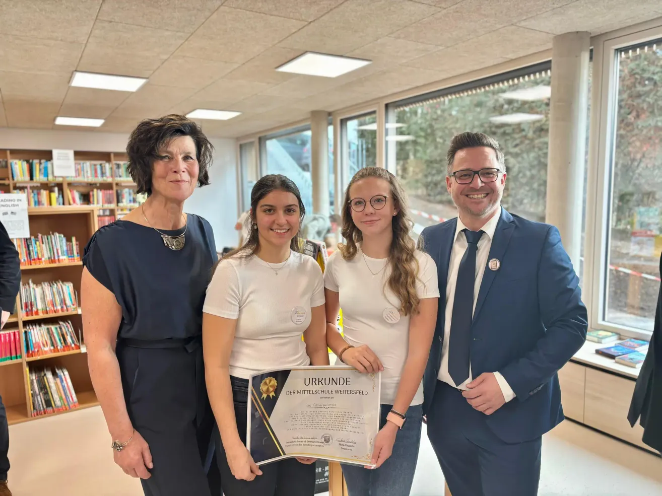 Zwei Frauen und zwei Männer stehen in einem Raum. Die Frauen halten ein Zertifikat und lächeln. Der Mann auf der rechten Seite trägt eine Brille und eine Krawatte. Sie stehen vor einer Glaswand.
