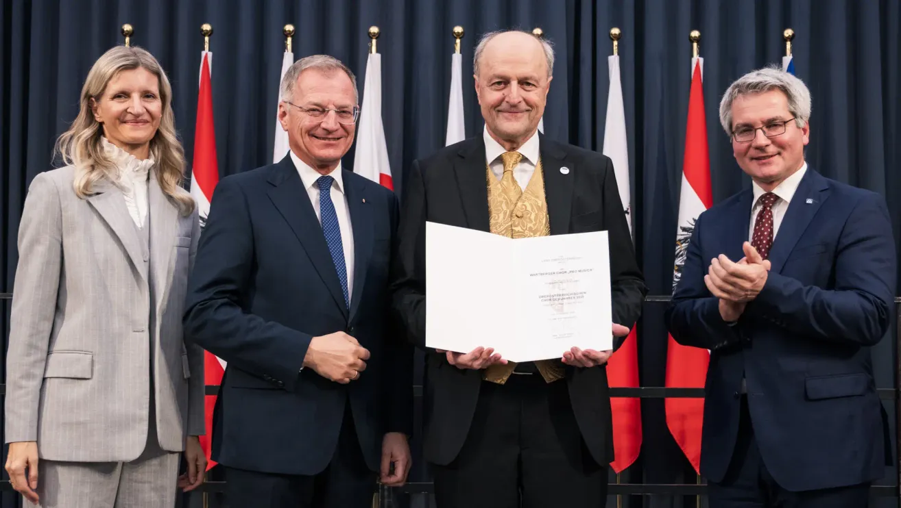 Zwei Männer in formeller Kleidung stehen vor einem Hintergrund mit Fahnen. Einer der Männer hält eine Urkunde, und beide lächeln.