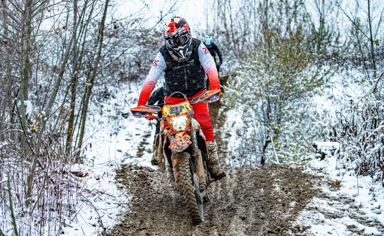 Ein Motocross-Fahrer fährt durch verschneiteres Gelände, ein weiterer Fahrer folgt. Sie tragen Helme und Schutzkleidung. Bäume umgeben sie, und der Boden ist schlammig.