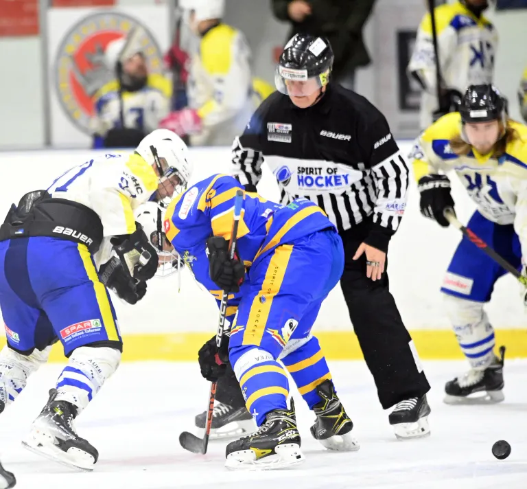 Ein Eishockeyspiel ist im Gange. Spieler in blauen und weißen Uniformen treten auf dem Eis an, während ein Schiedsrichter in der Nähe steht. Ein Spieler in Blau ist gebeugt und hält einen Eishockeyschläger.