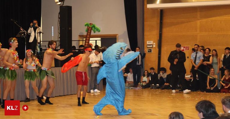 Eine Gruppe von Menschen befindet sich auf einer Tanzfläche mit einer Person in einem Haikostüm und zwei anderen in roten Shorts und Leis. Zuschauer sitzen hinter ihnen auf dem Boden.