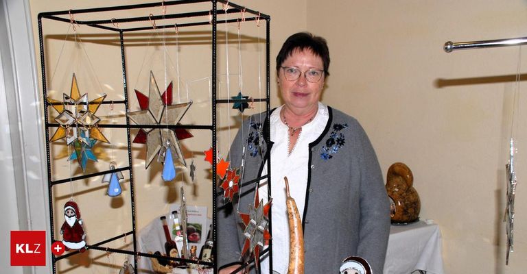 Eine ältere Frau mit Brille steht neben einer Ausstellung von Glasschmuck und Kunsthandwerk. Die Ornamente umfassen Sterne und Engel. Sie trägt eine graue Strickjacke mit einem weißen Hemd darunter.