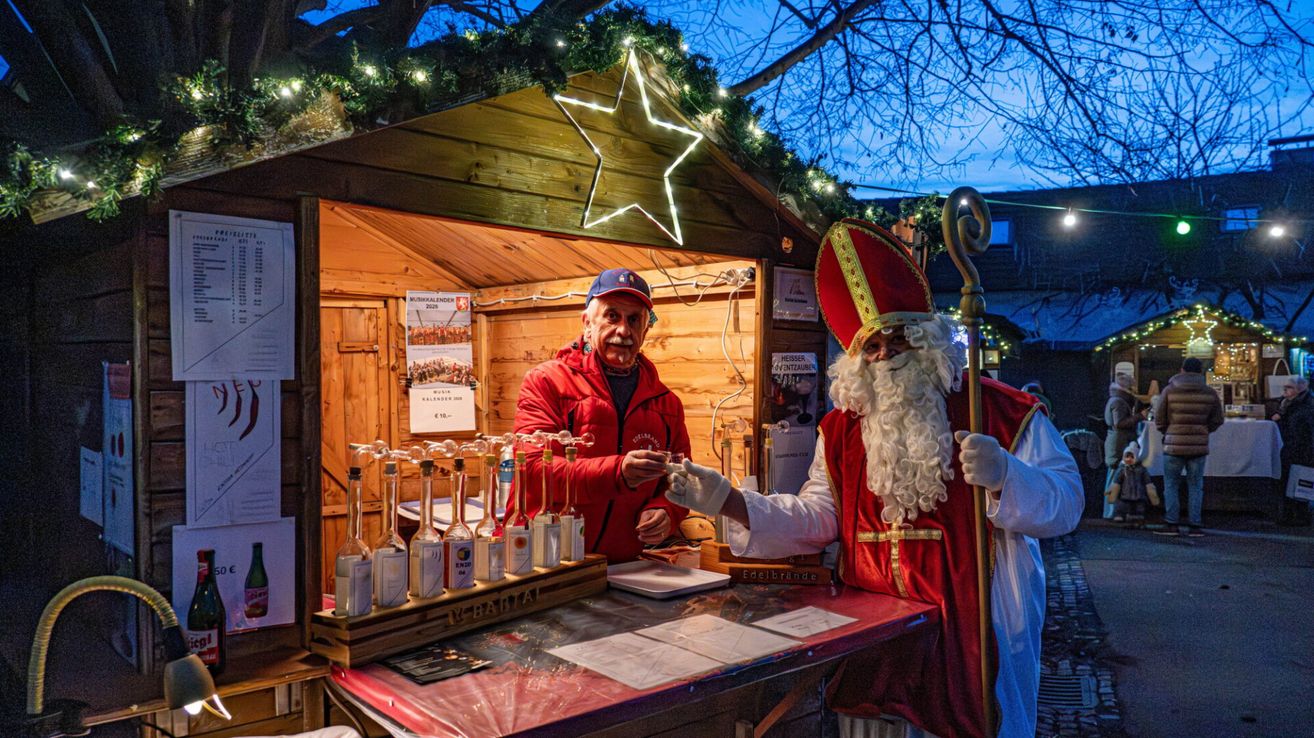 Ein Mann, der als Weihnachtsmann verkleidet ist, hält einen Stab und reicht einem Mann in einem roten Mantel bei einem Weihnachtsmarktstand ein Glas.