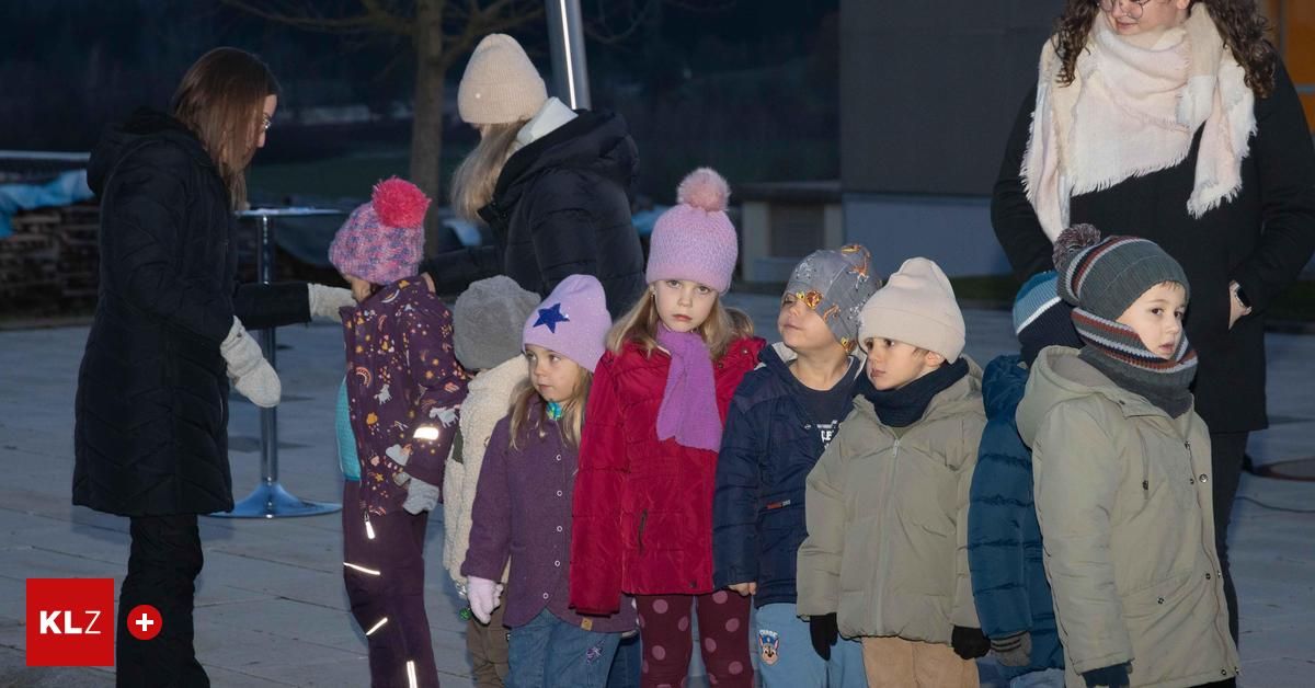 Eine Gruppe von Kindern in Winterkleidung steht bei Nacht draußen. Sie schauen nach vorne und tragen Mützen mit Bommeln. Ein Erwachsener steht hinter ihnen.