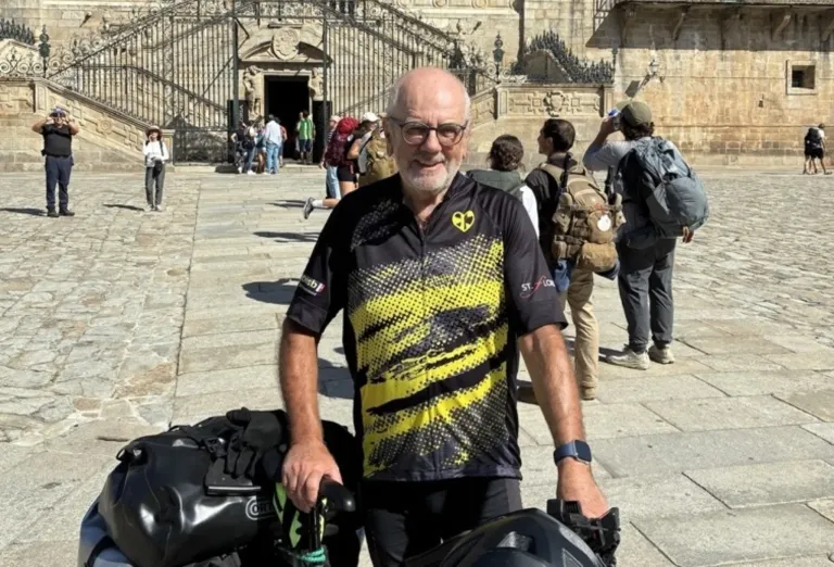 Ein älterer Mann in einem gelben und schwarzen Fahrradtrikot steht lächelnd vor einem Steingebäude. Mehrere Personen mit Rucksäcken gehen hinter ihm.