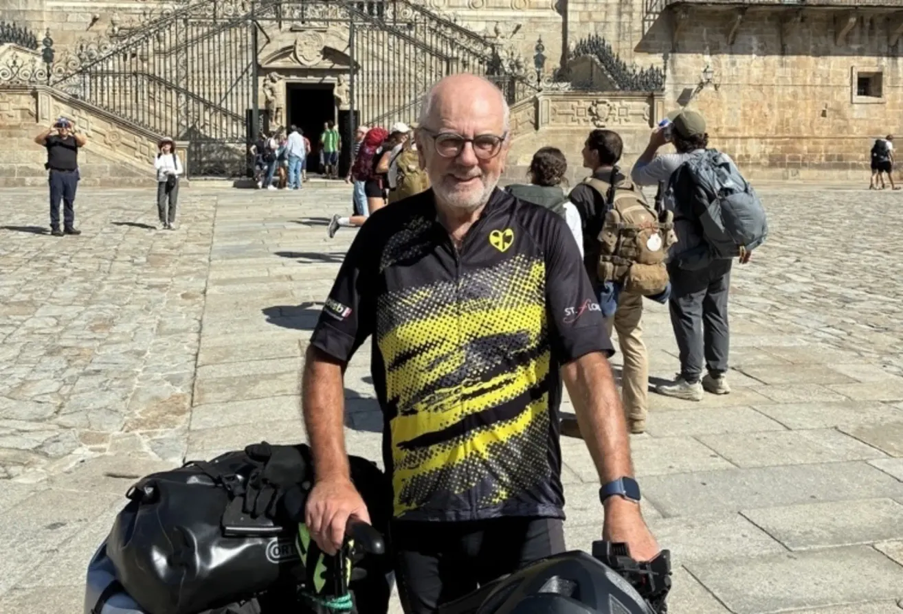 Ein älterer Mann in einem gelben und schwarzen Fahrradtrikot steht lächelnd vor einem Steingebäude. Mehrere Personen mit Rucksäcken gehen hinter ihm.