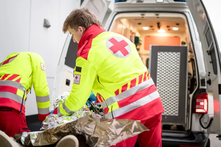 Ein Sanitäter in einer gelben Jacke mit rotem Kreuz auf der Vorderseite steht neben einem offenen Krankenwagen, möglicherweise bereit, einen Patienten zu transportieren.
