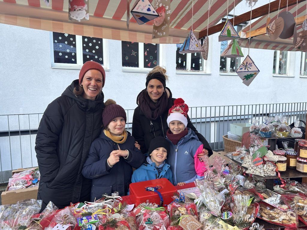 Eine Familie posiert lächelnd für ein Foto auf einem festlichen Markt. Sie sind von verschiedenen Weihnachtsdekorationen und Geschenken umgeben, mit einer roten Box im Vordergrund.