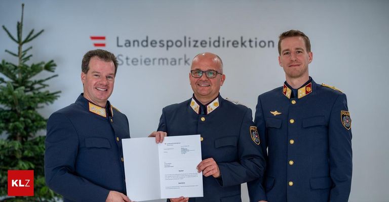 Drei Männer in Uniform stehen zusammen und halten ein Zertifikat. Der Mann in der Mitte trägt eine Brille. Hinter ihnen steht ein Schild mit der Aufschrift 'Landespolizeidirektion Steiermark'.