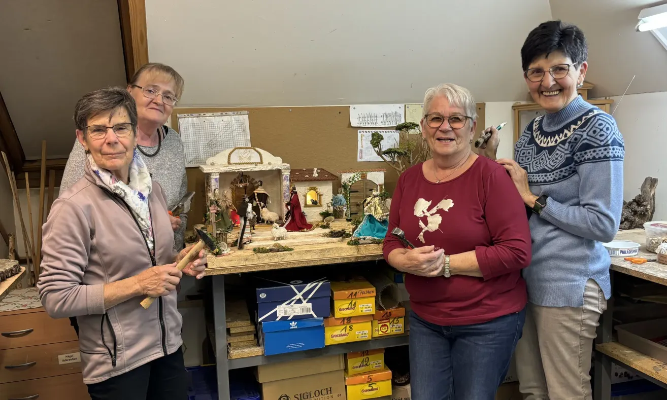 Eine Gruppe älterer Frauen lächelt und steht um einen Tisch mit einer kleinen Krippe darauf. Eine Frau hält einen Hammer.