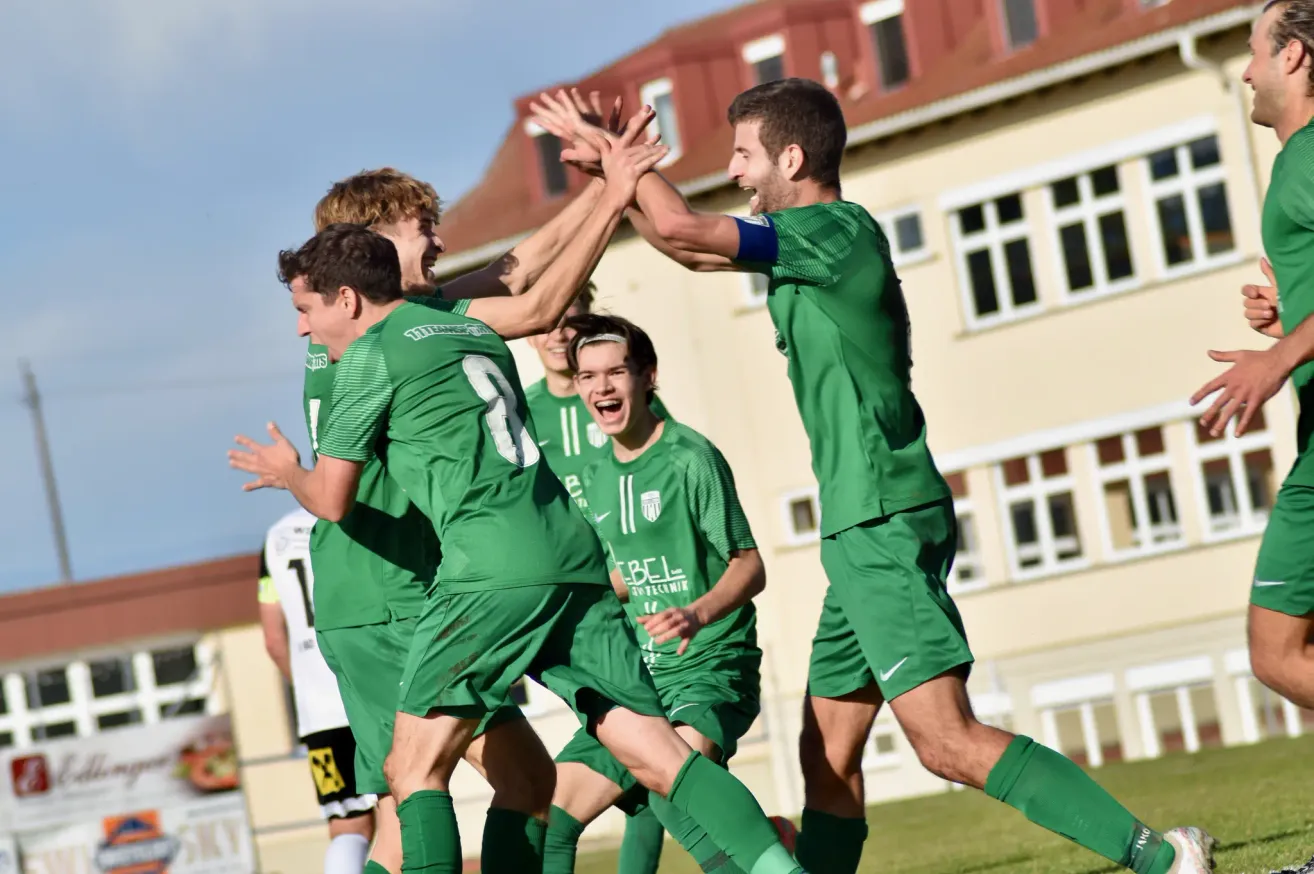 Eine Gruppe von Fußballspielern in grünen Trikots feiert ein Tor, wobei ein Spieler die Nummer 8 trägt. Sie stehen vor einem Gebäude mit roten Dächern und weißen Fenstern.