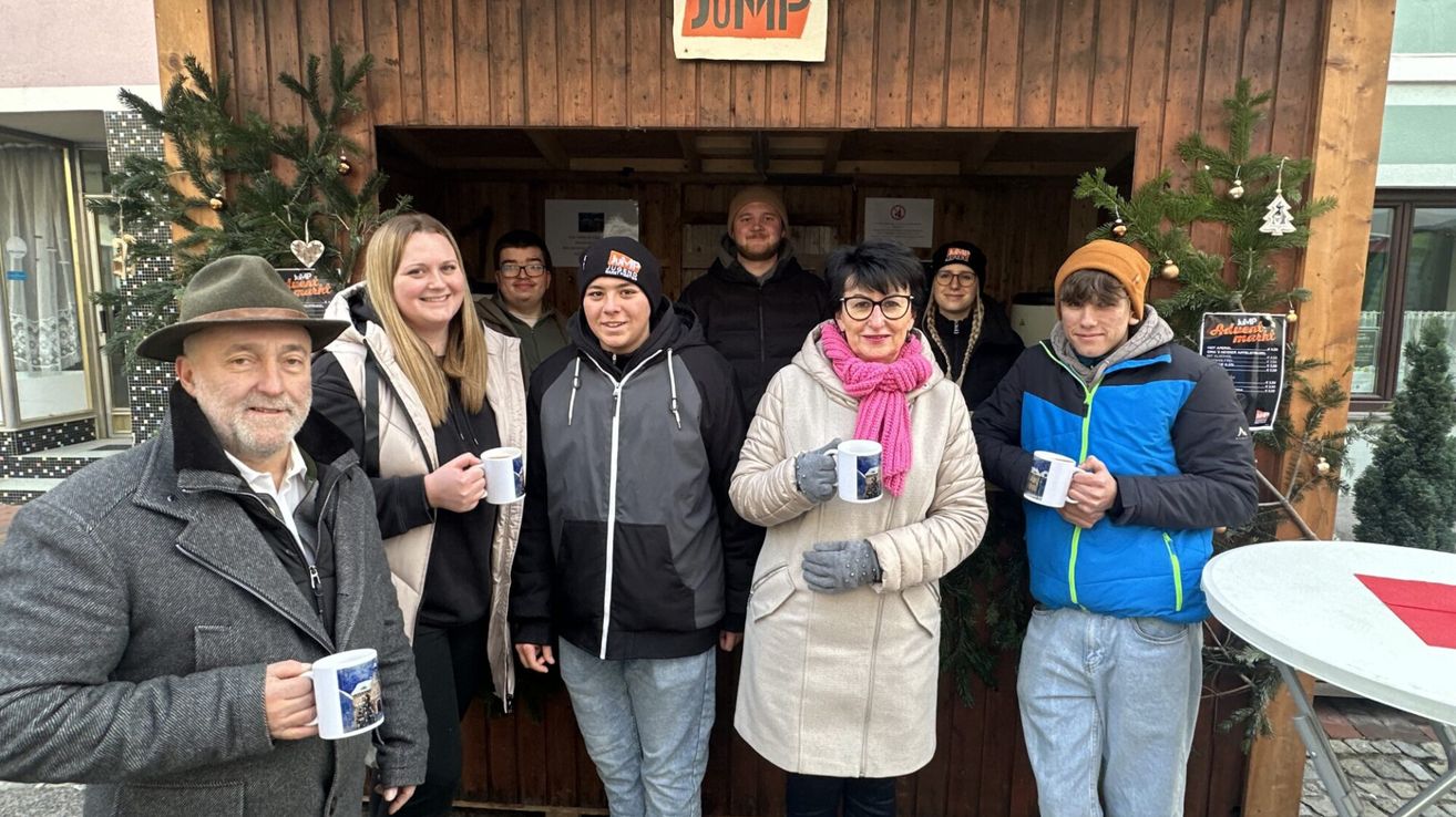 Eine Gruppe von Menschen steht vor einer Holzstruktur mit einem Schild, auf dem JUMP steht. Sie tragen alle Winterkleidung und halten Tassen. Es gibt Weihnachtsdekorationen an der Wand.