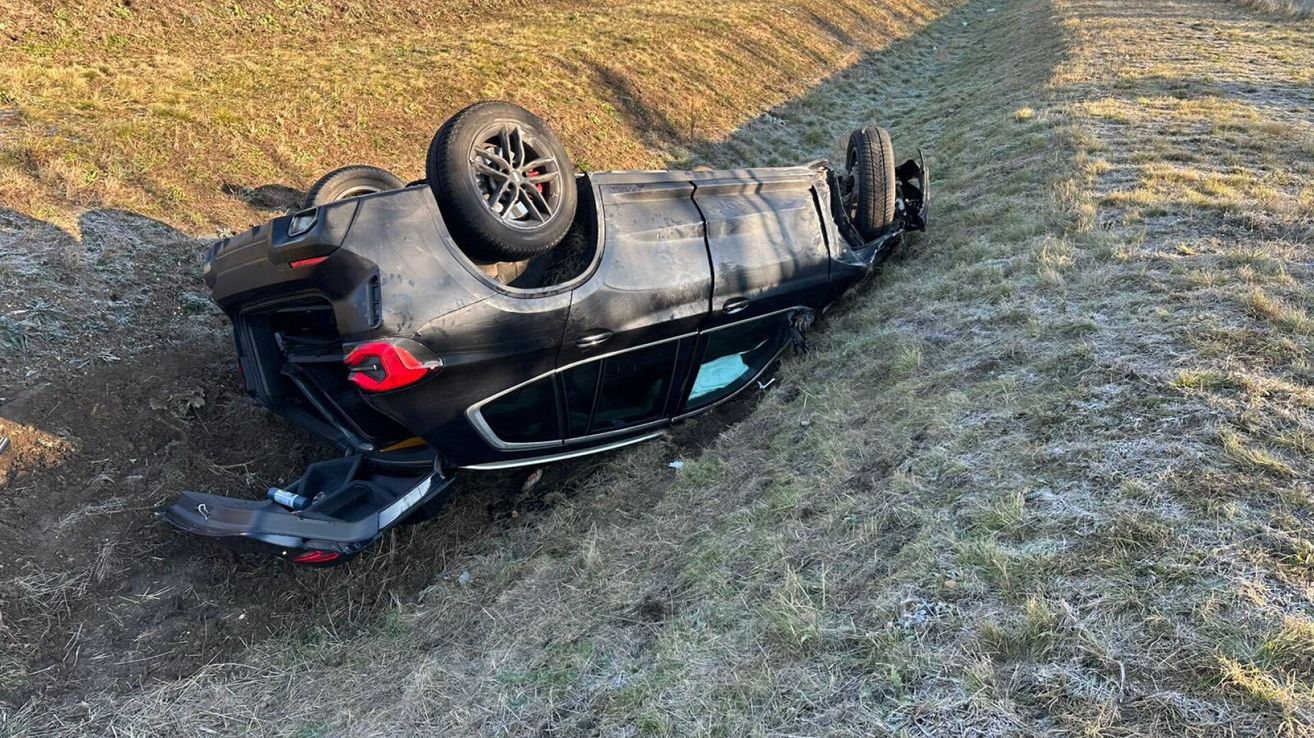 Ein schwarzer SUV liegt auf einem Grashang umgedreht, mit den Reifen in der Luft. Die Front des Autos ist kaputt und der Kofferraum ist offen.