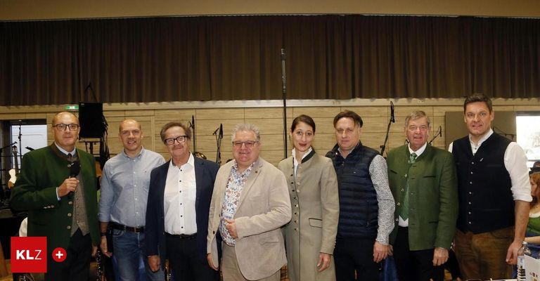 Sechs Personen, vier Männer und zwei Frauen, gekleidet in formeller Kleidung, posieren für ein Foto vor einer Bühne mit Mikrofonen.