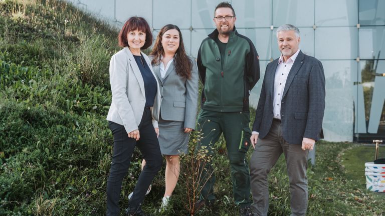 Vier Personen in formeller Kleidung stehen zusammen draußen. Zwei Frauen und zwei Männer, alle lächeln und posieren für ein Foto.