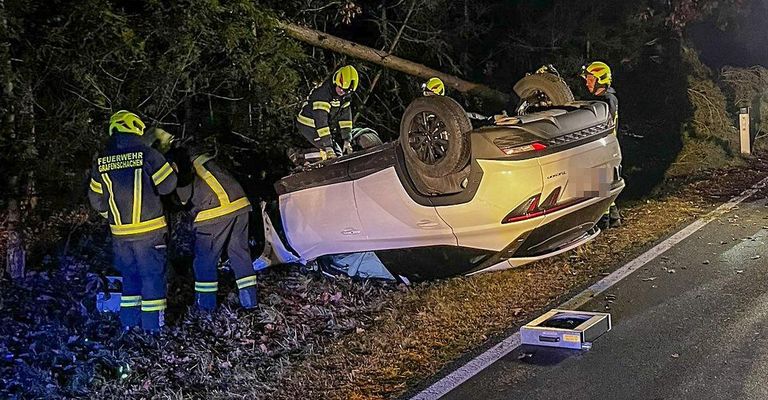 Feuerwehrleute kümmern sich um ein umgestürztes Auto am Straßenrand. Das Auto liegt auf der Seite, und die Feuerwehrleute bereiten sich darauf vor, die Insassen zu retten.