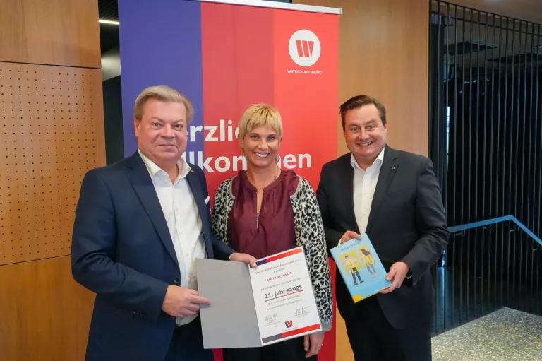 Drei Menschen stehen für ein Foto zusammen, zwei Männer und eine Frau. Sie halten Urkunden und ein Buch. Ein Banner hinter ihnen hat Text und ein Logo.