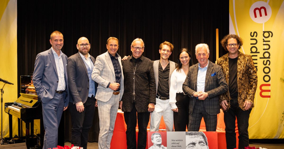 Eine Gruppe von Fachleuten steht für ein Foto auf der Bühne mit einem Hintergrund. Sie tragen formelle Kleidung, und eine Person hat einen Namensschild. Sie stehen eng beieinander und lächeln für die Kamera.