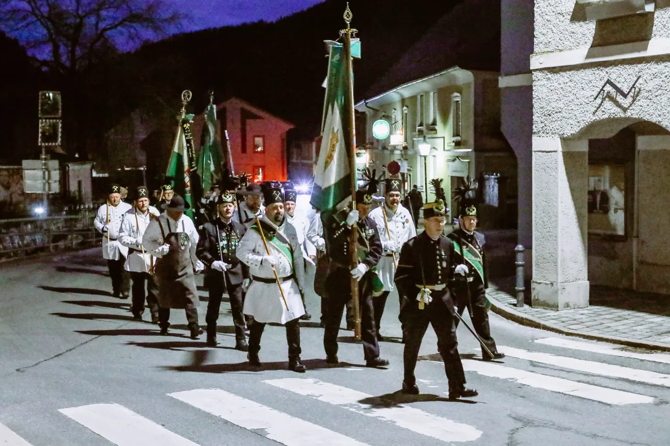 Ein nächtlicher Zug mit uniformierten Personen, die Fahnen und Stäbe halten, auf einer Stadtstraße. Gebäude und eine grüne Straßenlaterne sind im Hintergrund zu sehen.