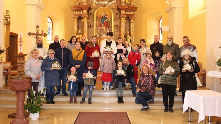 Eine Gruppe von Erwachsenen und Kindern posiert vor einem Altar in einer Kirche, jeder hält eine kleine Struktur, möglicherweise ein Modellhaus, in einer festlichen Atmosphäre.