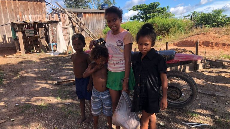 Drei Kinder stehen vor einem rostigen Motorrad und einem Haus. Ein Mädchen hält eine Plastiktüte und die anderen beiden Jungen sehen zu.