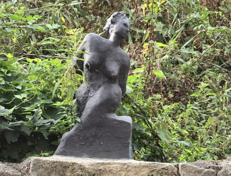 Eine Bronzestatue einer Frau steht im Freien. Sie ist nackt und scheint in sitzender Position zu sein. Die Statue ist von Grünpflanzen umgeben.