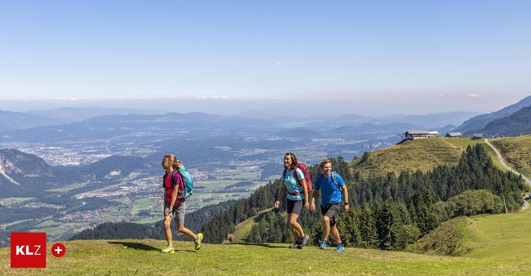 Drei Wanderer gehen auf einem grasbewachsenen Hügel, tragen Rucksäcke und Turnschuhe. Der Hintergrund zeigt einen Panoramablick auf Berge, eine Stadt und einen Fluss.