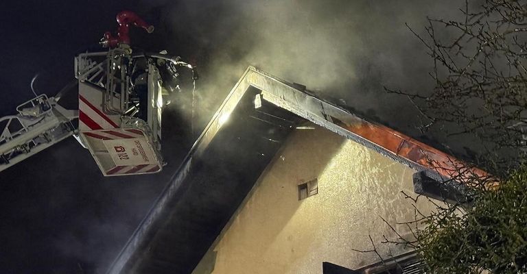 Feuerwehrleute sind auf einer Leiter und spritzen Wasser auf ein brennendes Haus in der Nacht. Das Dach zeigt Schäden, und Rauch steigt vom Gebäude auf.