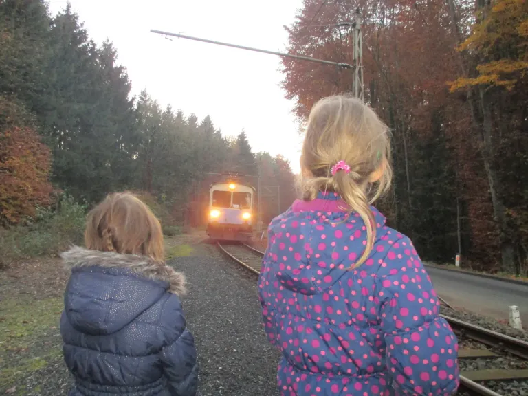 Zwei Mädchen in Winterjacken beobachten einen herannahenden Zug auf einem Gleis, umgeben von Bäumen im Hintergrund.