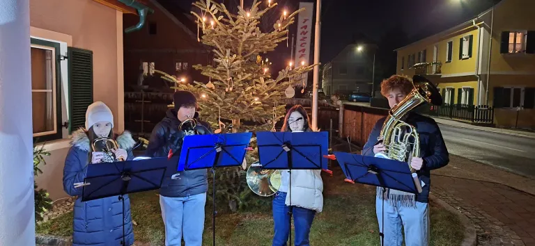 Drei Musiker stehen vor einem geschmückten Weihnachtsbaum und spielen Blechblasinstrumente. Die Szene ist bei Nacht, mit einem Gebäude und einem Auto im Hintergrund sichtbar.