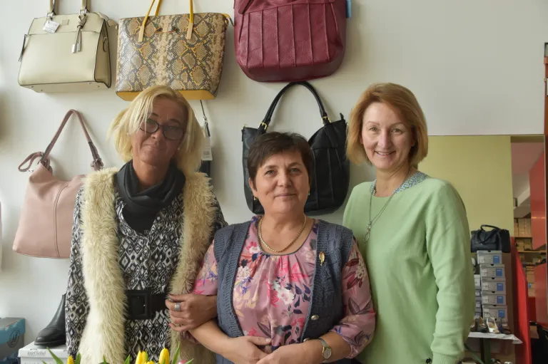 Three women stand together in front of a wall adorned with various handbags. Each woman wears a different outfit and accessory, and they appear to be posing for a photo.