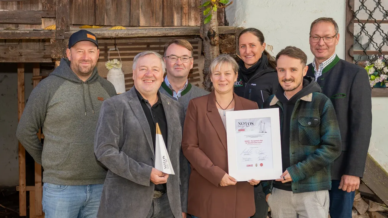 Fünf Personen stehen nebeneinander und lächeln für ein Foto. Eine Frau hält ein Zertifikat. Sie sind draußen mit einer Holzwand und einem Baum im Hintergrund.