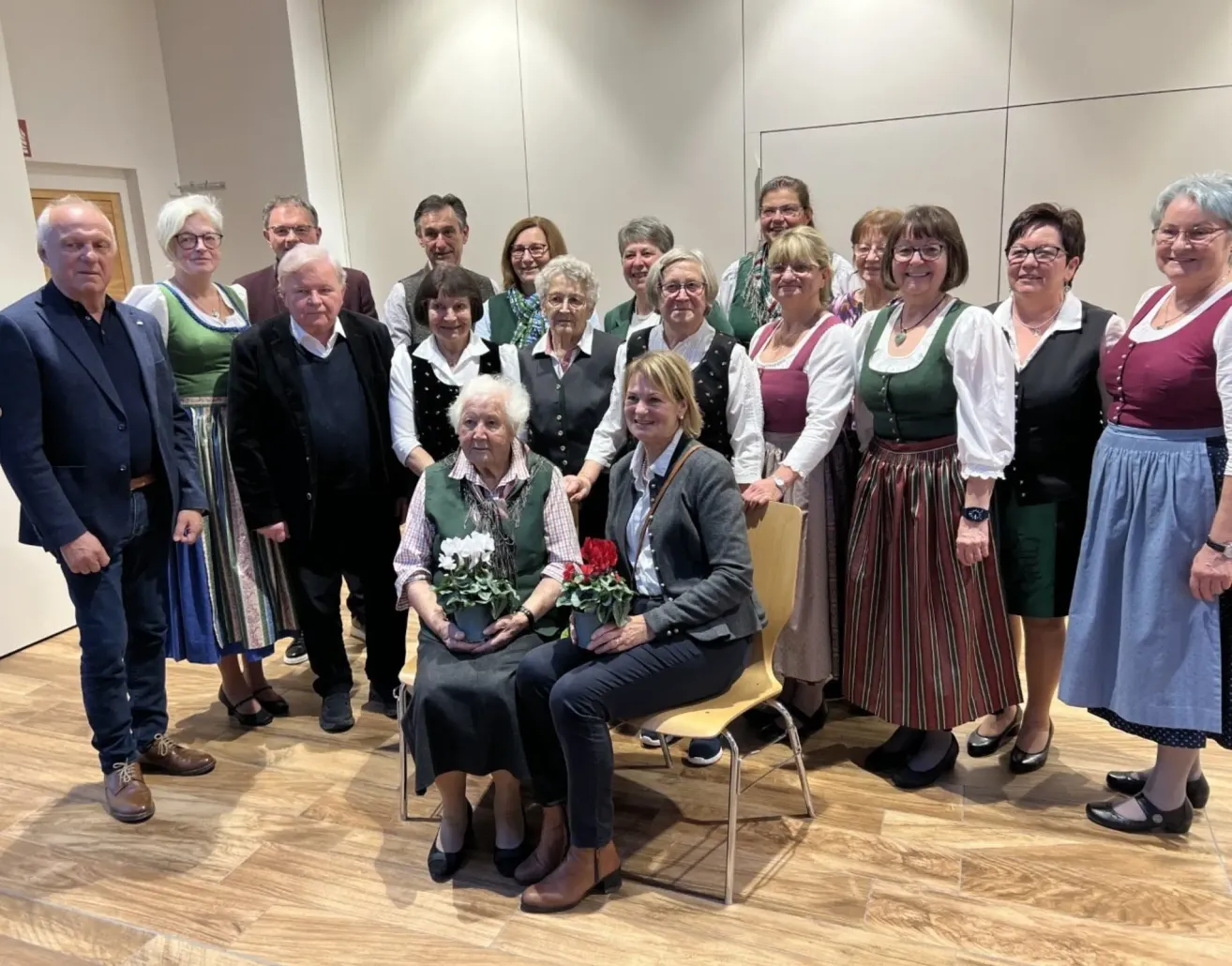Eine Gruppe von Erwachsenen in traditioneller Kleidung posiert für ein Foto in einem Raum, wobei zwei Frauen im Vordergrund sitzen und Blumen halten.