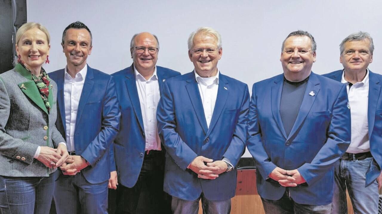 Vier Männer in blauen Anzügen stehen lächelnd zusammen, zwei von ihnen tragen Brillen. Sie scheinen sich in einer professionellen Umgebung zu befinden.