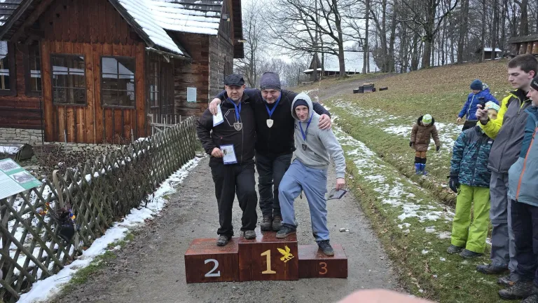 Drei Personen stehen auf einem Podest, zwei Männer mit Medaillen um den Hals und ein Junge, der ein Papier in der Hand hält. Sie befinden sich in einem verschneiten Bereich.