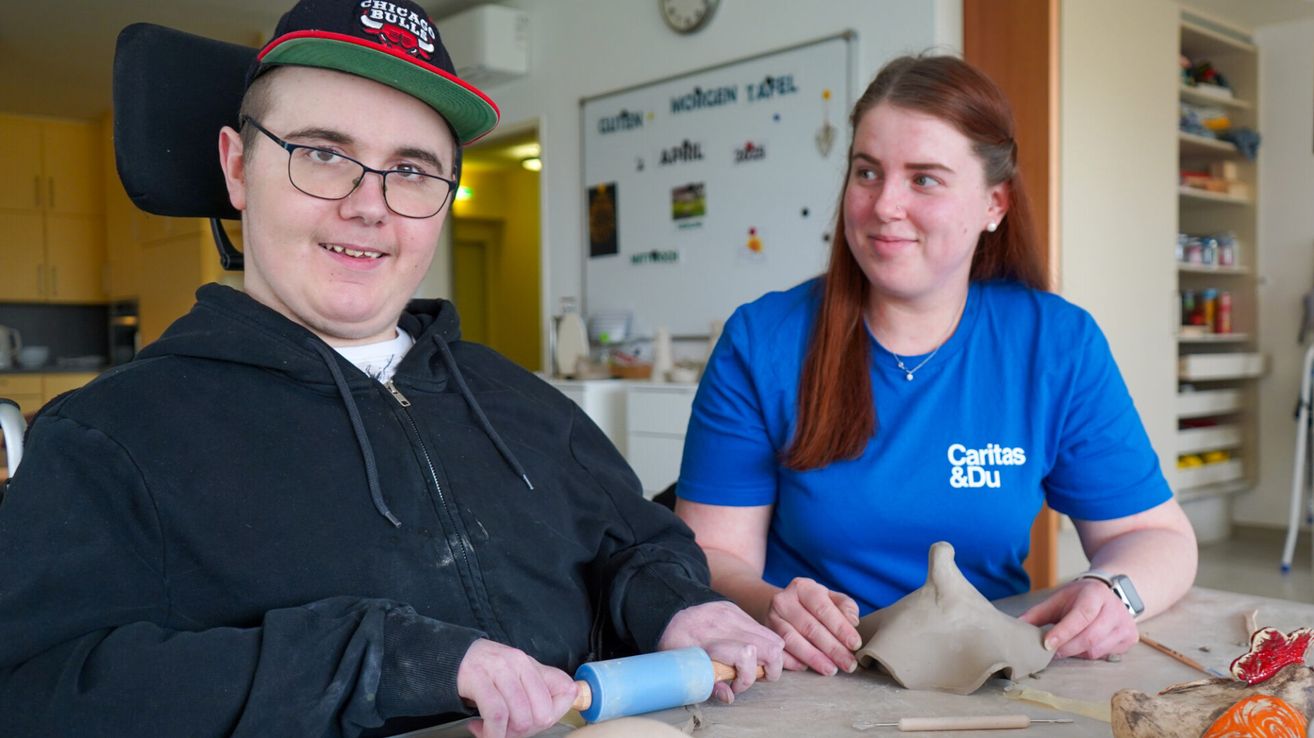 Ein Mann und eine Frau arbeiten in einem Raum an Töpferwaren. Der Mann trägt eine Chicago Bulls Kappe und eine Brille. Die Frau trägt ein blaues T-Shirt mit Caritas & Du. Sie lächeln beide und scheinen die Aktivität zu genießen.