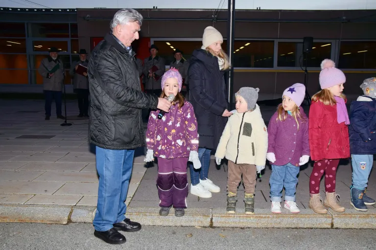 Ein erwachsener Mann hält ein Mikrofon und spricht zu einer Gruppe von Kindern. Die Kinder sind in Winterkleidung gekleidet und stehen auf dem Bürgersteig.