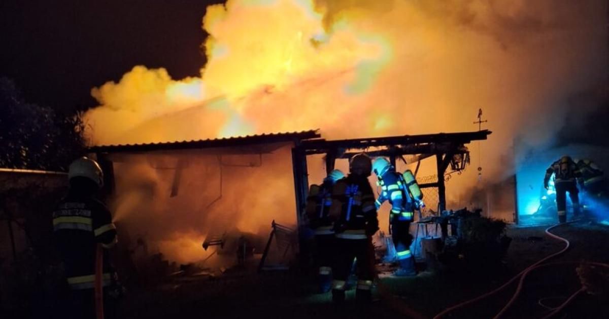 Drei Feuerwehrleute stehen vor einem brennenden Gebäude, beleuchtet vom Feuerschein, mit Rauch, der um sie herum aufsteigt.