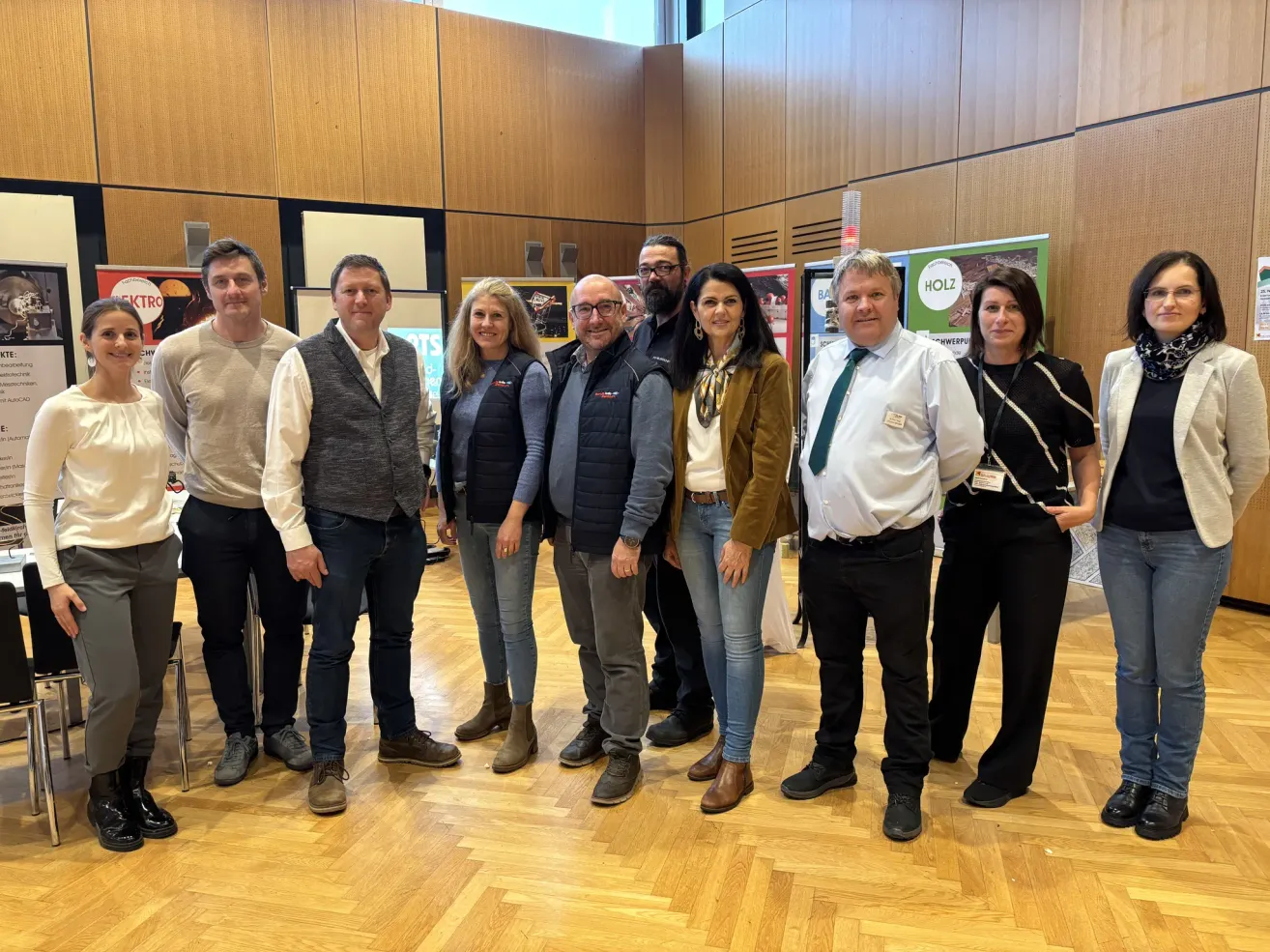 Eine Gruppe von Menschen steht in einem Raum mit Holzböden. Sie posieren für ein Foto. Sie tragen formelle Kleidung. Hinter ihnen befinden sich Holzwände mit Postern. Der Raum hat eine warme und gemütliche Atmosphäre.