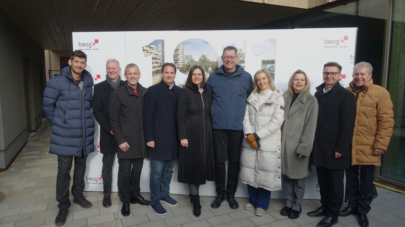 Eine Gruppe von sieben Erwachsenen steht vor einer großen weißen Wand mit der Nummer 100. Sie alle lächeln und scheinen für ein Foto zu posieren.