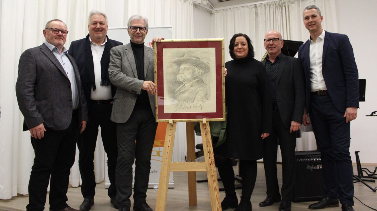 Vier Personen stehen lächelnd für ein Foto zusammen. Ein Mann hält ein gerahmtes Porträt. Hinter ihnen hängt ein weißer Vorhang an der Wand.